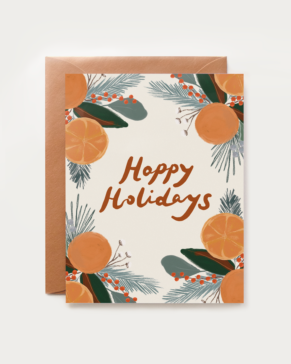 A holiday greeting card with a copper envelope behind it, featuring the words “Happy Holidays” in brown script and surrounded by illustrated citrus slices, green leaves, and blue-gray pine needles
