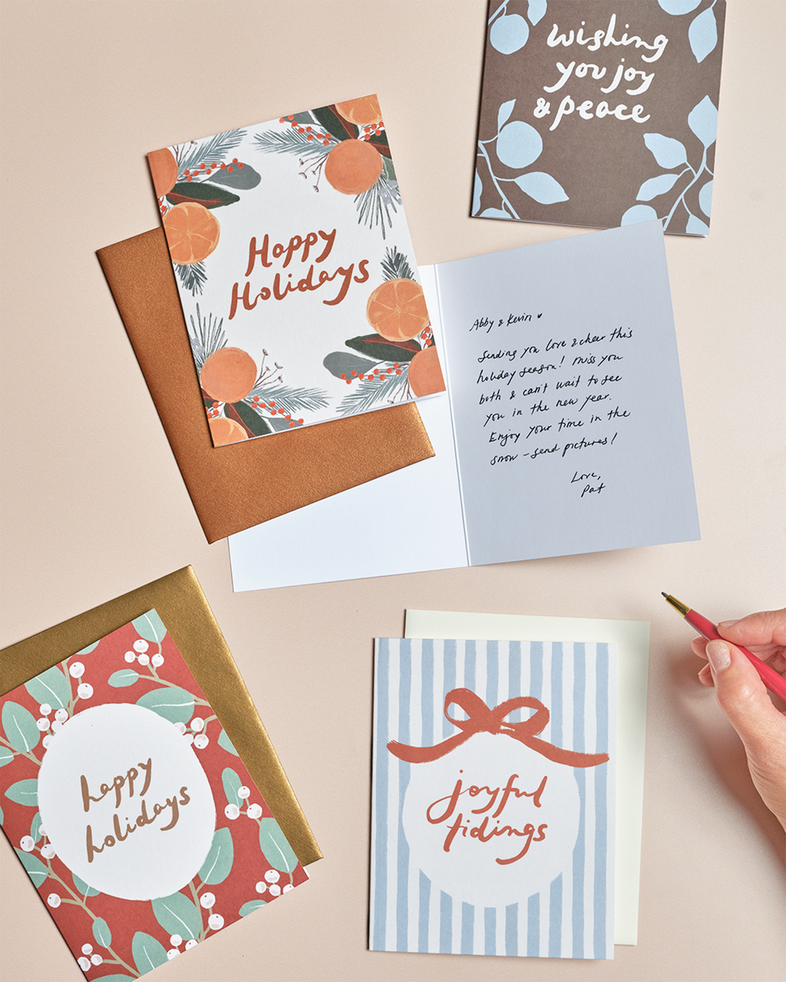 A styled flat-lay of multiple holiday greeting cards and envelopes on a warm beige background, with a hand holding a pink pencil at the lower right. Cards feature festive designs including orange citrus, red berries, blue leaves, striped ribbons, and handwritten holiday messages.